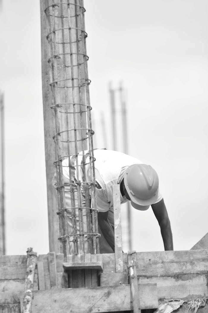 about-07 Grayscale image of a construction worker bending over a steel frame, taken outdoors.