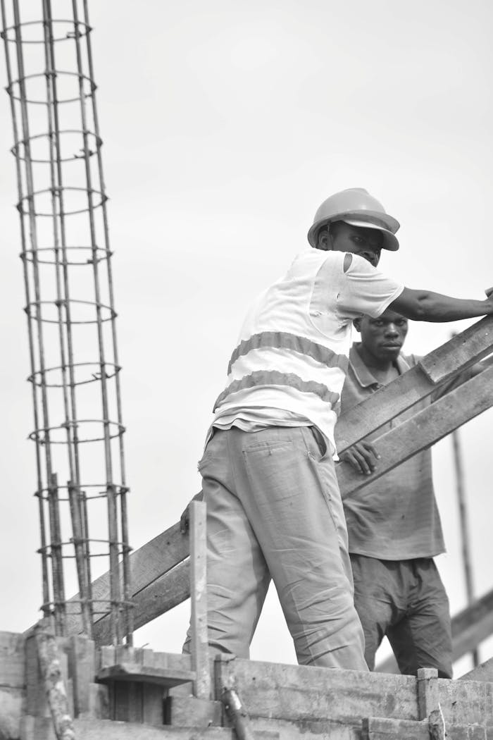 Black and white image of two construction workers wearing safety gear and helmets at a building site.