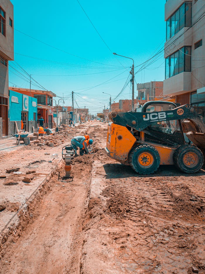 Busy urban road construction scene with workers and construction machinery under a bright blue sky.